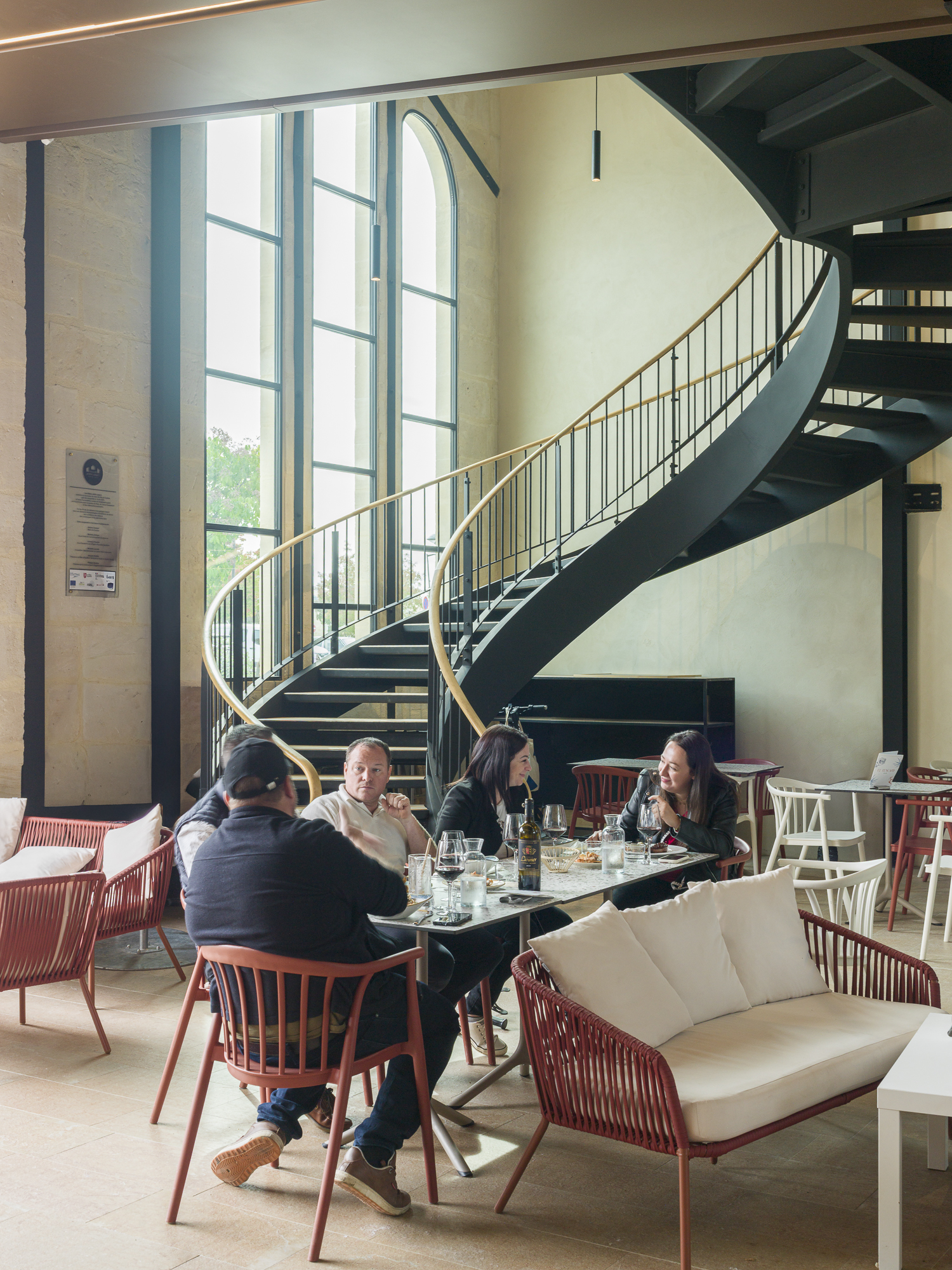 Prolongement intérieur de l'espace public sous le porche principal. Vue sur l'escalier monumental et les fenêtres existantes rabaissées qui offrent de nouvelles perspectives entre le marché et la place. / Nicolas Bouriette - Agence Bouriette Vaconsin - Réhabilitation des Halles Justin Luquot, Coutras, 2025.