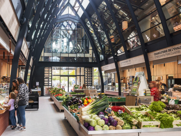 Jour de marché sous la verrière bioclimatique. Vue de l'espace de vente principal en rez-de-chaussée vers la place Ernest Barraud. / Nicolas Bouriette - Agence Bouriette Vaconsin - Réhabilitation des Halles Justin Luquot, Coutras, 2025.